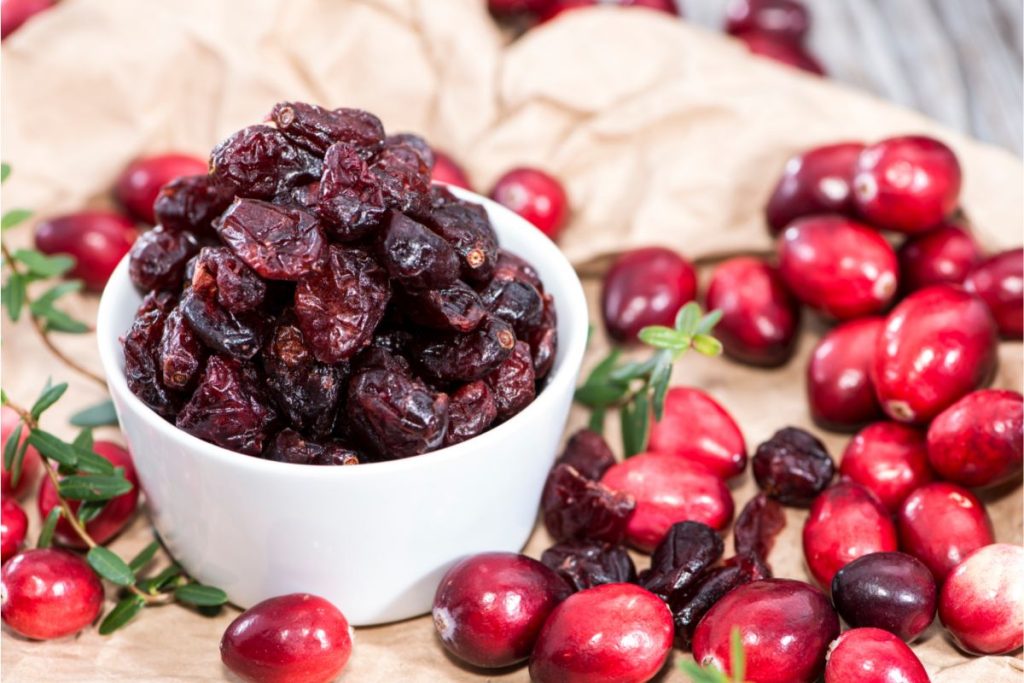 Dried cranberries in bowl, next to fresh cranberries scattered on a table.