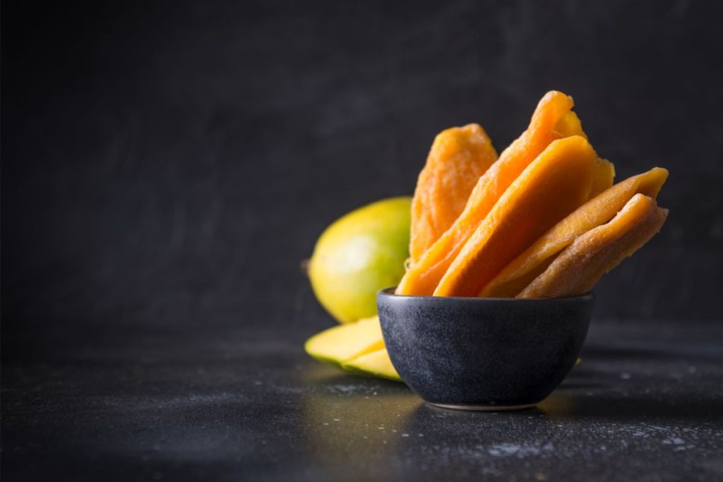 Dried mango chips in a bowl with fresh mangos in the background