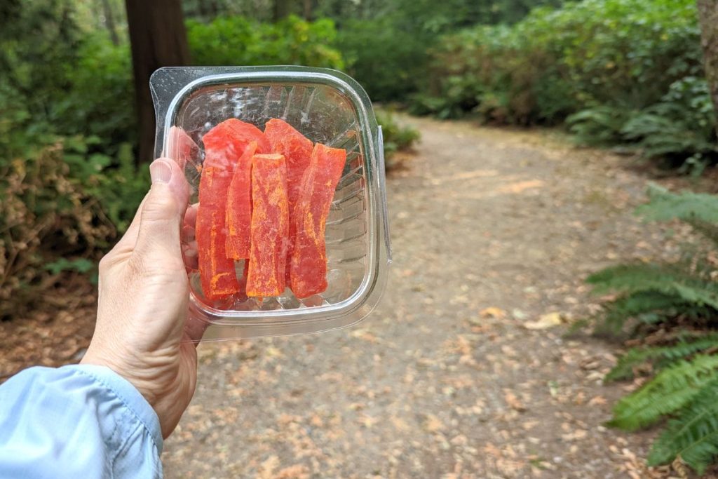 Dehydrated papaya slices held by a woman in a plastic clear container outdoors on a hiking trail