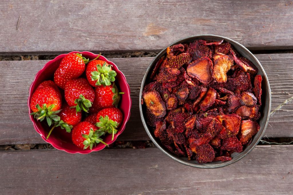 A bowl of fresh strawberries next to a bowl of sun-dried strawberry slices