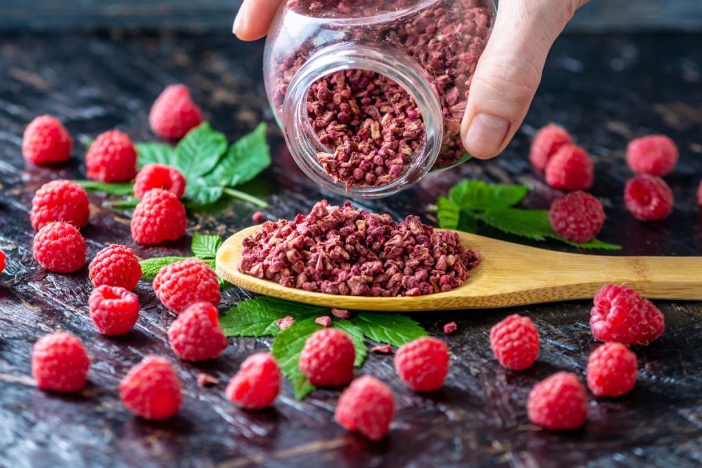 Woman shaking a jar full of ground freeze-dried raspberries into a wooden spoon surrounded by fresh raspberries