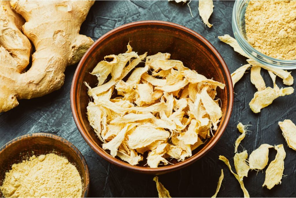 Bowl of dried ginger root next to ginger powder and a fresh ginger root