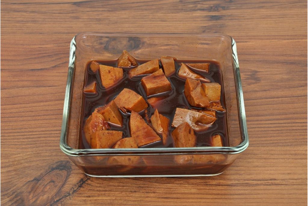 Tofu chunks sitting in a dark brown marinade in a glass container